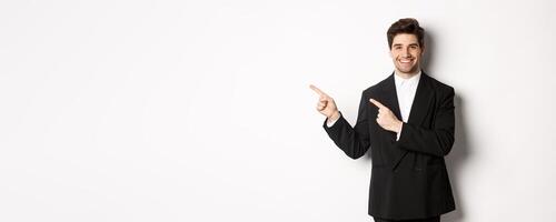 Portrait of successful, handsome businessman in suit, pointing fingers at upper left corner and smiling, showing advertisement, standing over white background photo