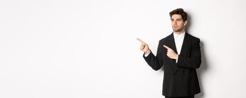 Portrait of serious handsome man in black suit, pointing and looking left at promo banner, showing advertisement on copy space, standing over white background photo