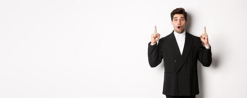 Portrait of surprised handsome businessman in black suit, saying wow and looking amazed, pointing fingers up at copy space, standing over white background photo