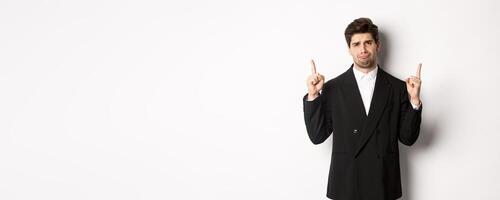Portrait of reluctant and sad handsome man, complaining and pointing fingers up at something bad, looking jealous, standing over white background photo