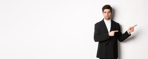 Image of handsome businessman in black suit, pointing fingers right and looking at camera, standing against white background photo