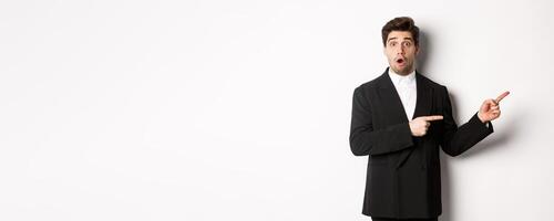Portrait of surprised handsome man with beard, wearing black suit, open mouth and pointing fingers right at advertisement, standing over white background photo