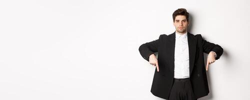 Image of handsome man dressed for formal party, wearing suit and pointing fingers down, showing advertisement or making announcement, standing over white background photo