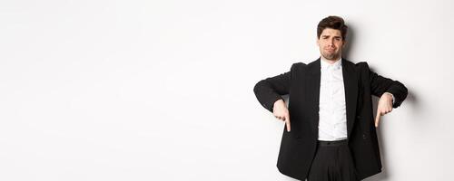 Portrait of skeptical and unamused bearded man, pointing fingers down with disappointed face, standing in suit and complaining photo