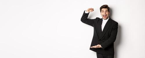 Portrait of excited handsome man in suit, shaping big object on copy space and smiling amazed, holding something, standing over white background photo