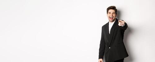 Concept of new year party, celebration and lifestyle. Portrait of handsome stylish man in black suit, smiling and pointing finger at camera, standing over white background photo