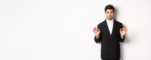 Concept of new year party, celebration and lifestyle. Portrait of confident good-looking man in black suit, showing okay sign and approve something, standing over white background photo