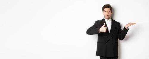 Portrait of hadnsome bearded man in formal suit, showing thumb-up and holding product in hand over white copy space, recommending product, standing over white background photo