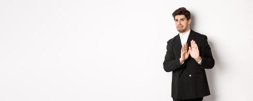 Image of stylish handsome man in black suit, cringe from something embarrassing, declining offer and showing stop gesture, standing against white background photo