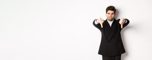 Portrait of skeptical and disappointed man in black suit, frowning upset, showing thumbs-down, dislike something bad, standing over white background photo