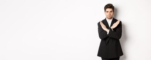 Portrait of serious and concerned man in black suit, showing stop gesture and frowning, making cross to prohibit or decline something, standing over white background photo