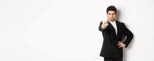 Portrait of serious handsome man in business suit, showing one finger to prohibit or decline something, telling to stop, disagree with you, standing over white background photo