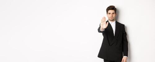 Portrait of serious handsome man in formal suit, extending hand to stop you, prohibit action, forbid and disagree with something, standing against white background photo