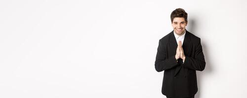 Portrait of handsome man in black suit, being grateful, saying thank you and bowing politely, smiling as holding hands together, expressing gratitude, standing over white background photo