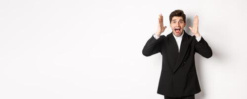 Frustrated and concerned man in black suit, screaming in panic and looking at something shocking, standing over white background photo