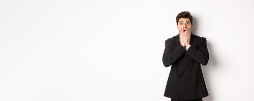 Image of concerned and scared man in black suit, gasping and looking alarmed at camera, standing over white background photo