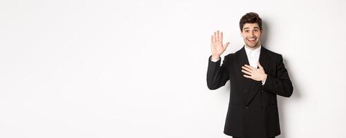 Image of handsome smiling guy telling the truth, raising on arm to promise something, standing over white background in black suit photo