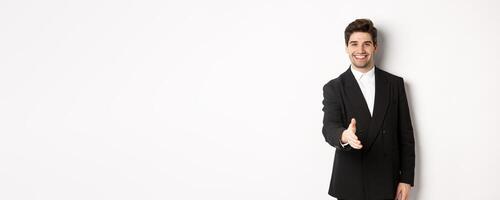 Portrait of handsome businessman in black suit, extending hand for handshake, greet business partners and smiling, welcome to company, standing over white background photo