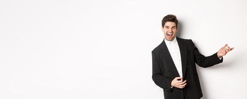 Portrait of happy man dancing at party, playing on invisible guitar and laughing, standing in black suit against white background photo