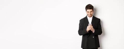 Portrait of handsome stylish businessman in black suit, writing a message, smiling and looking at smartphone, standing over white background photo
