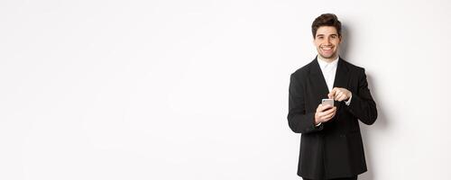Image of handsome businessman in suit showing you app on smartphone, pointing finger at mobile screen and smiling, standing over white background photo