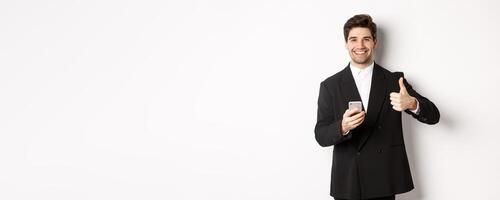 Handsome guy in trendy suit making compliment, showing thumbs-up while using mobile phone, recommending an app or online shop, standing over white background photo