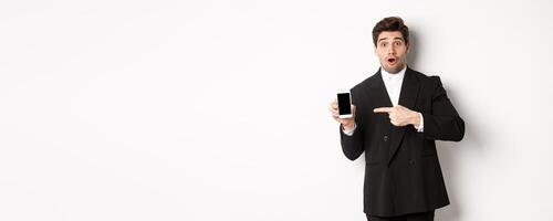 Portrait of attractive businessman in black suit, looking surprised and pointing finger at smartphone sreen, standing over white background photo