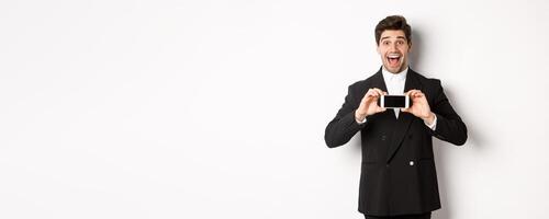 Image of cheerful, handsome man in black suit, showing smarthone screen and looking amazed, standing against white background photo