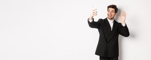 Image of handsome man in suit, having call and waving hand at smartphone camera, recording , greeting someone, standing against white background photo