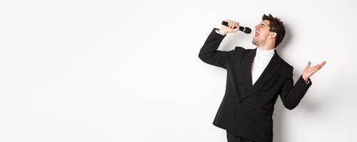 Portrait of handsome man singing a song with passion, standing in black suit, holding microphone and performning, posing over white background photo