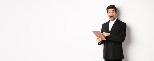 Portrait of amazed handsome businessman, wearing trendy suit, showing something cool on digital tablet, standing against white background photo
