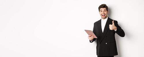 Portrait of handsome businessman in suit, showing thumb-up and making a compliment, praising something on digital tablet, standing over white background photo