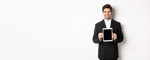 Portrait of handsome businessman in trendy suit, showing digital tablet screen and smiling, standing against white background photo
