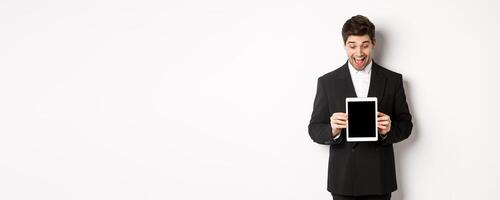 Portrait of amazed handsome man in black suit, showing digital tablet screen with excitement, standing over white background photo
