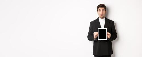 Image of surprised handsome man in black suit, showing digital tablet screen and looking amazed, standing against white background photo