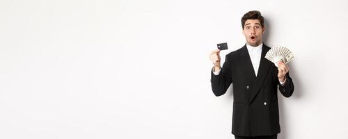 Portrait of surprised handsome man i suit, showing credit card with money, standing against white background photo