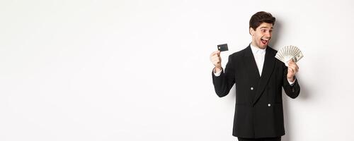 Image of attractive businessman in black suit, rejoicing, showing credit card and looking at money, standing against white background photo