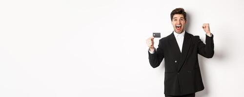 Portrait of excited handsome businessman in suit, rejoicing and showing credit card, standing against white background photo