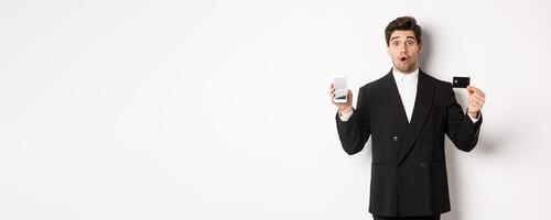 Image of handsome businessman in black suit, looking amazed and showing credit card with mobile phone screen, standing against white background photo
