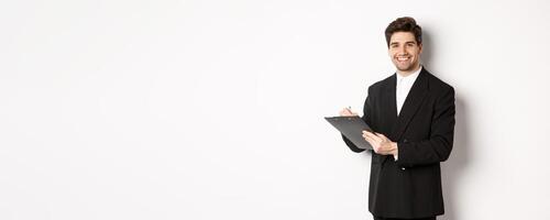 Portrait of confident businessman in black suit, signing documents and smiling, standing happy against white background photo