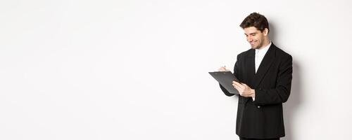 Image of handsome businessman in suit signing documents, looking at clipboard and smiling, standing against white background photo