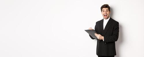 Portrait of handsome businessman in black suit, looking amazed while signing documents on clipboard, standing against white background photo
