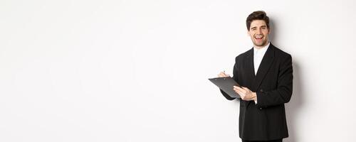 Image of handsome, successful boss in black suit, winking and smiling while signing documents, standing against white background photo