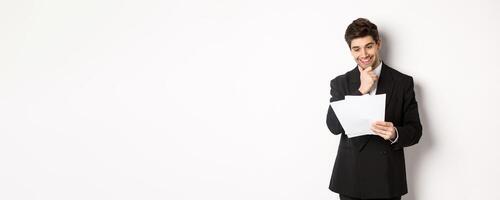 Image of handsome businessman in black suit, looking pleased at documents, reading report and smiling, standing against white background photo