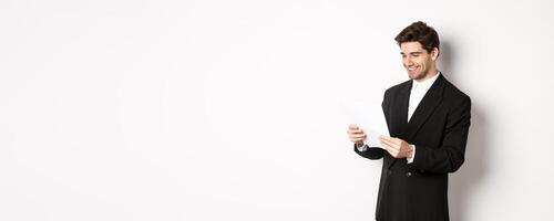 Image of attractive businessman in black suit, reading document and smiling, working on report, standing against white background photo