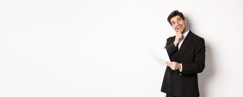 Image of thoughtful, handsome businessman in black suit, holding document and looking at upper right corner, thinking and smiling, standing against white background photo