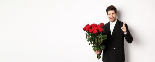 Image of handsome romantic guy in black suit, holding bouquet of roses and pointing at camera, congratulating with holiday, standing against white background photo