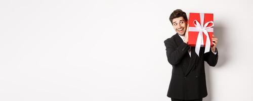 Concept of christmas holidays, celebration and lifestyle. Image of excited man enjoying new year, shaking gift box to guess what inside, standing against white background photo