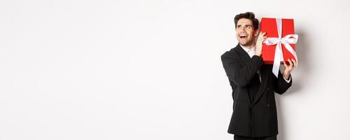 Concept of christmas holidays, celebration and lifestyle. Image of excited man enjoying new year, shaking gift box to guess what inside, standing against white background photo