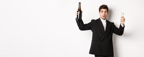 Concept of holidays, party and celebration. Image of handsome man in stylish suit, dancing with bottle of champagne, drinking on new year, standing over white background photo